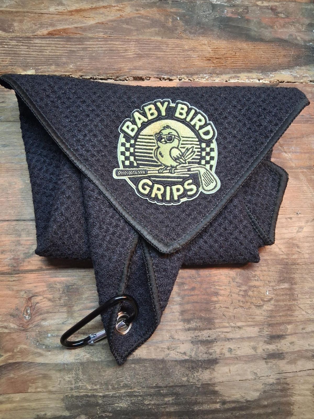 A black waffle golf towel with a green Baby Bird Grips patch and a carabiner