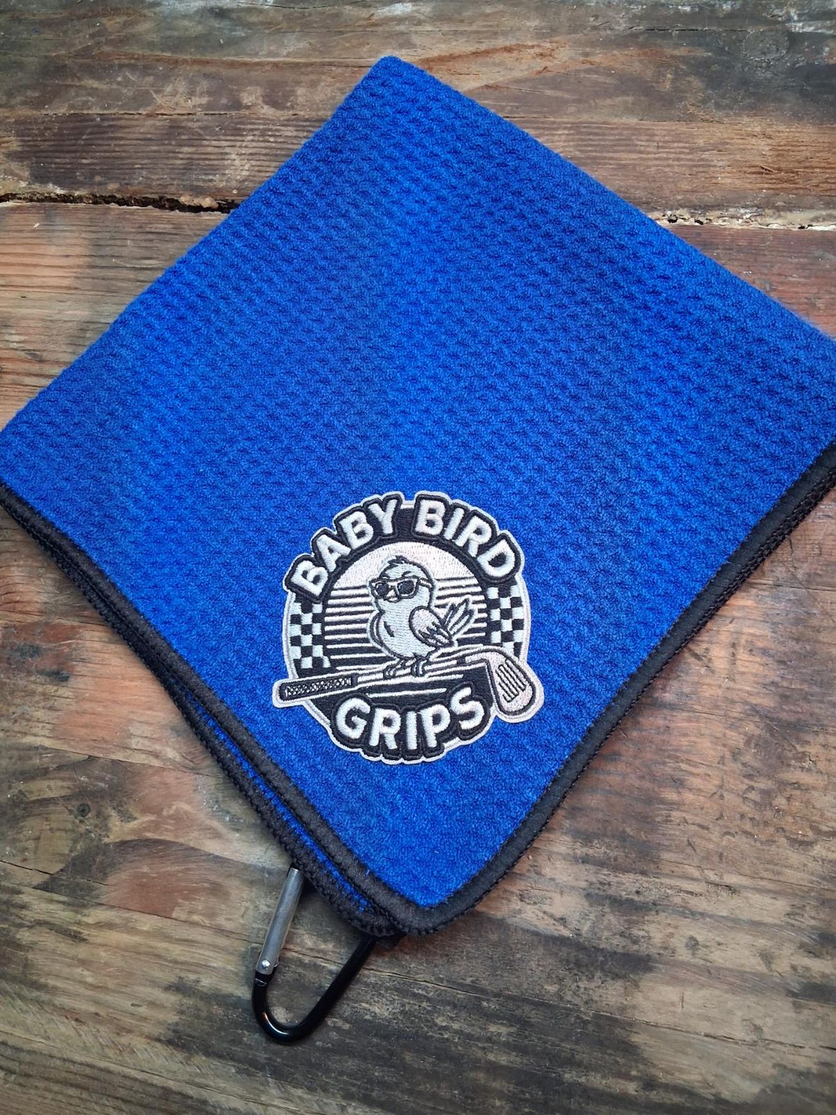 A blue waffle golf towel with a gray Baby Bird Grips patch and a carabiner